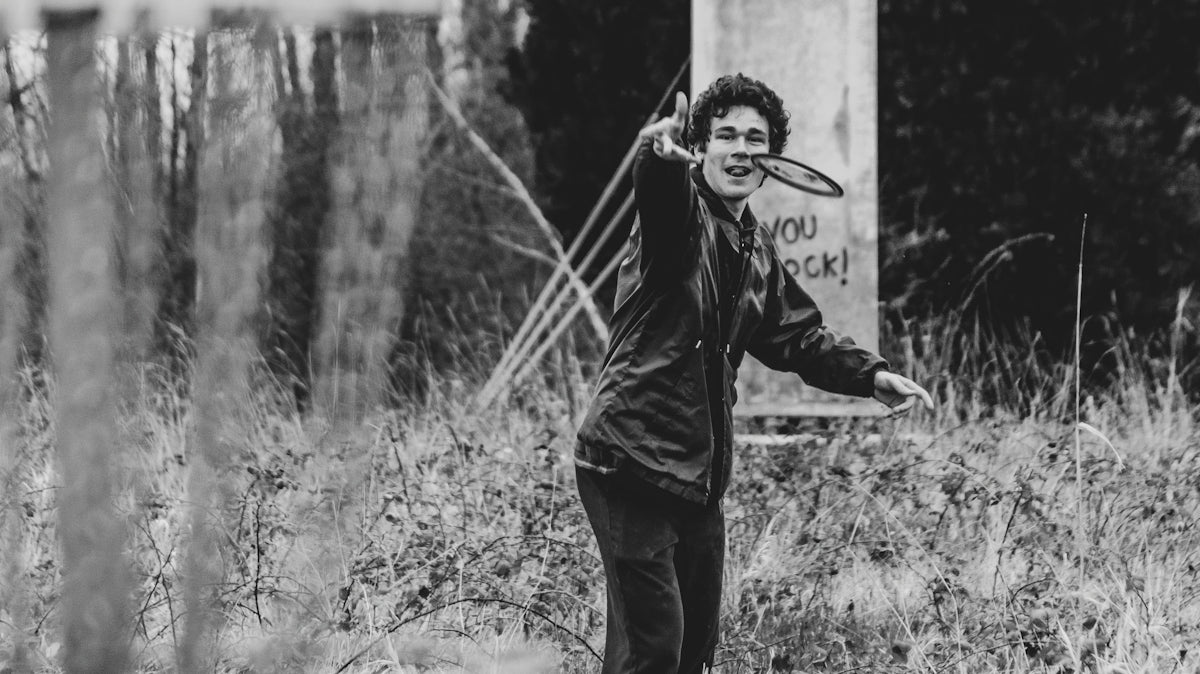 man in black jacket and pants standing beside concrete post in grayscale photography