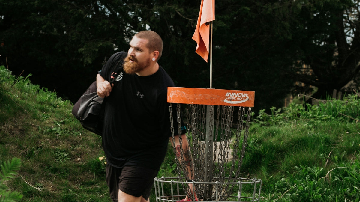 Man walking past a disc golf basket with a bag.