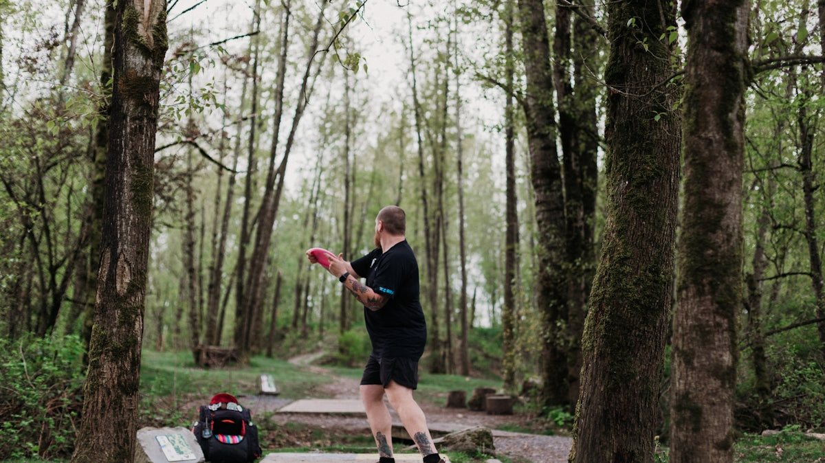 Man throwing a disc golf disc in a wooded park.